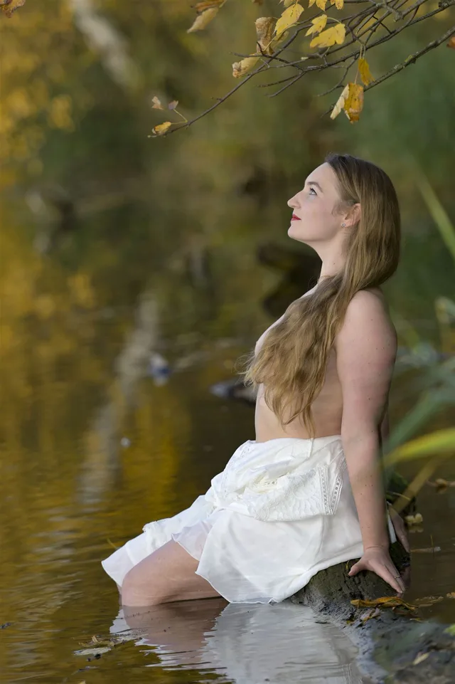 Frau mit weißem Stoff sitzt unter einem Ast am Wasser und blickt nach oben.