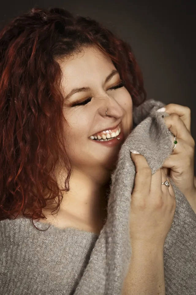 Lachende Frau mit geschlossenen Augen, die ihren grauen Pullover an die Wange hält.